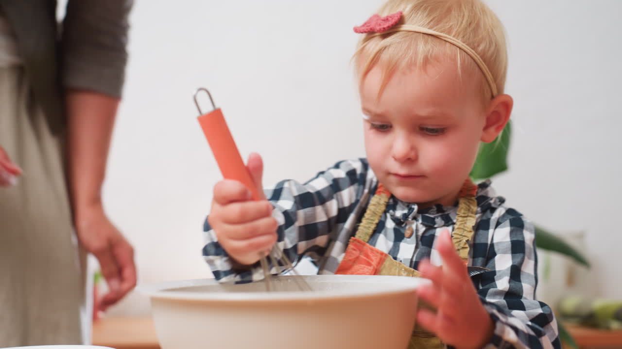 Bambina finisce di setacciare la farina mentre la madre toglie il setaccio, preparandosi a darle la frusta per mescolare durante l'attività di preparazione al forno, concentrandosi sull'apprendimento in cucina e sulla collaborazione genitore-figlio