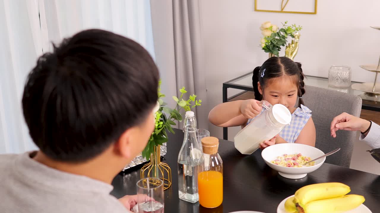 A young girl pours milk into cereal, assisted by her mother, while her father watches at a breakfast table
