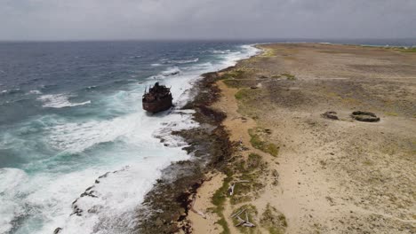 Aerial-fast-orbit-over-the-rusty-wreck-standing-on-top-of-the-reef-with-waves-crashing-and-the-lighthouse-in-the-back