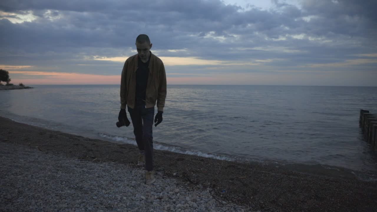 Man Holding Camera Walking And Leaving The Beach During Sunset By The Calm Ocean In Canada. - wide shot