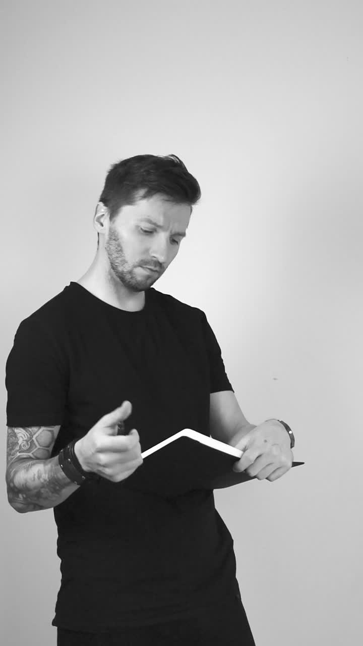 Grayscale footage of white tattooed short bearded and haired man writing something down in his notebook or journal. After writing lifts the pen up to his chin and begins to think and recollect on it.