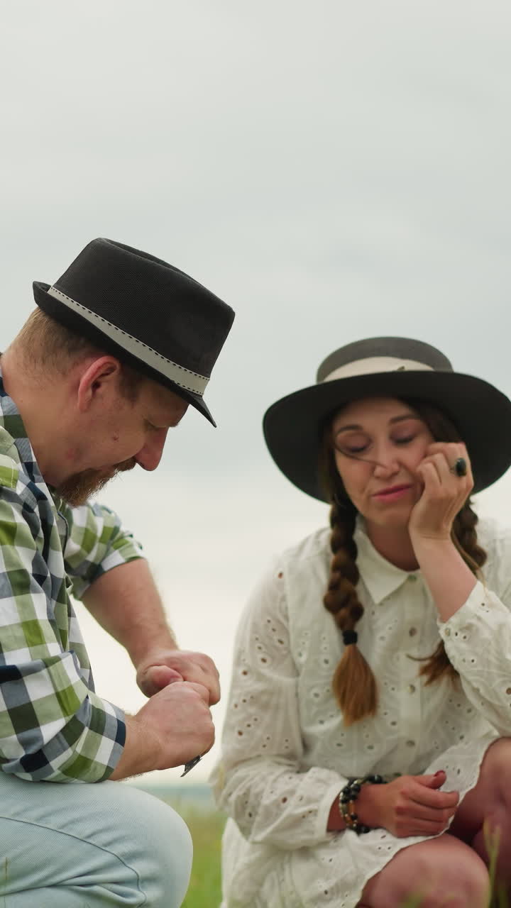 un hombre con una camisa a cuadros y un sombrero se esfuerza por abrir un tubo de pintura mientras una mujer con un vestido blanco y sombrero mira y sacude la cabeza con incredulidad