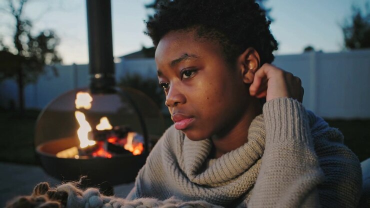 Chiminea fire flickering, teen adjusting hair and wrapping turtleneck on patio at dusk, reflecting
