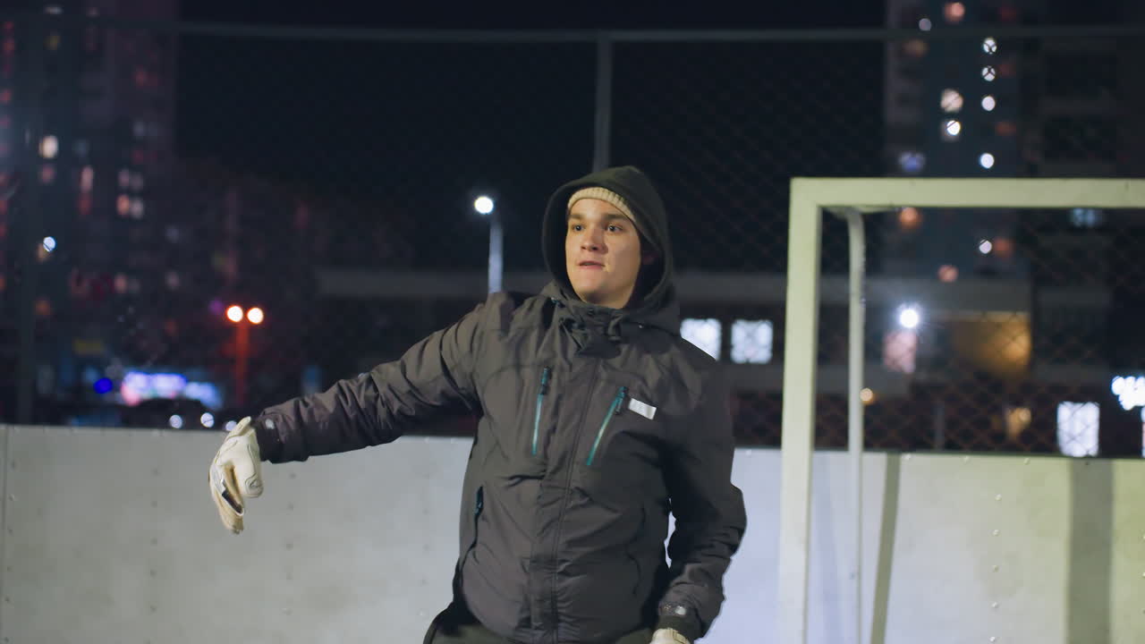 골키퍼가 골포스트 근처의 야간 연습 중에 축구공을 던지며, 뒤에 벽에 그림자가 여 있으며, 도시 배경의 조명된 주거 건물로 여 있습니다.