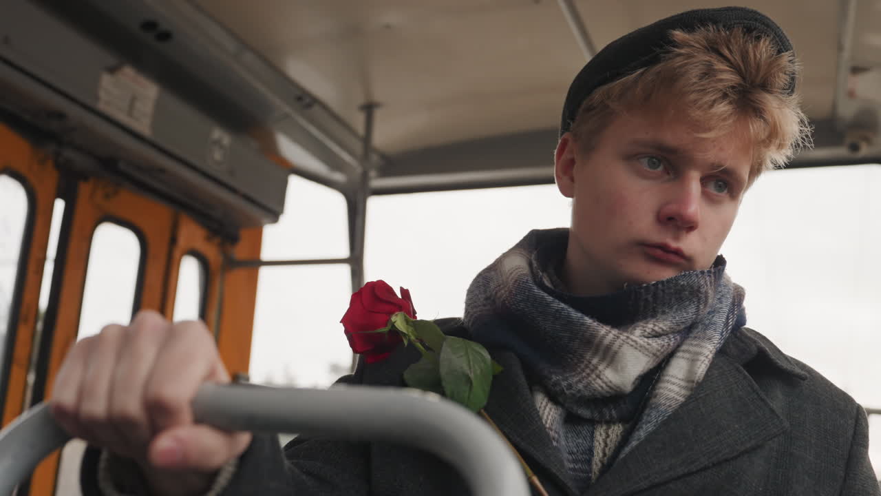 Nachdenklicher weißer Songwriter in der Straßenbahn, Rose. Junger weißer Musiker/Kreativer hält eine Rose, komponiert eine Melodie im Kopf, vorbeiziehende Stadt vor beschlagenem Fenster, Wollschal und Beanie, Nahaufnahme im Porträtformat, weich.