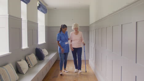 senior woman at home using walking stick being helped by female care worker in uniform