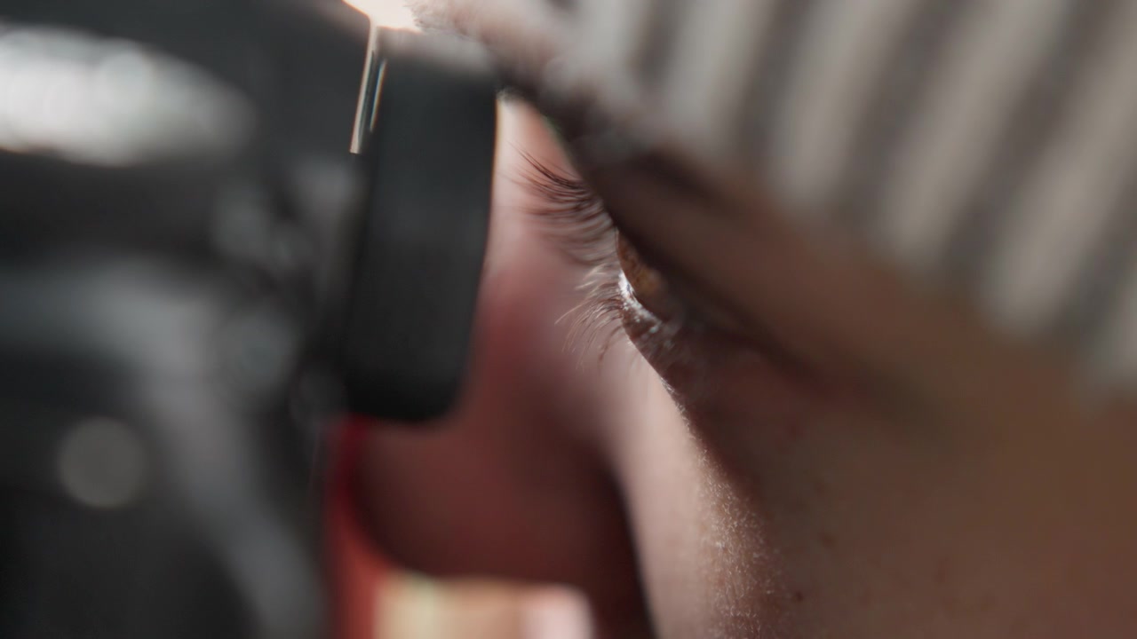 Tight eye portrait through camera, focused photographer peering through eyecup, soft skin tones, beanie shadow, concentrated composition, cinematic grain, natural lighting, creative direction, shallow