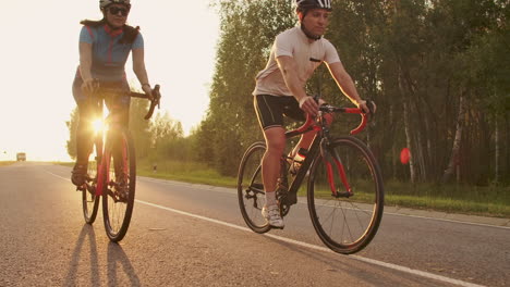 A-man-and-a-woman-on-bicycles-ride-down-the-road-at-sunset-together-in-slow-motion.-The-couple-travels-by-Bicycle.-Sports-Cycling-helmets.