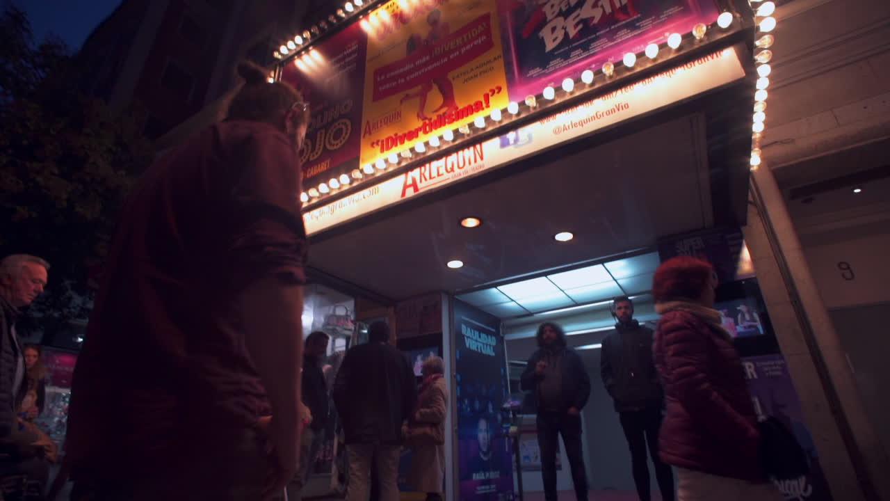 Panning low rotation around people walking in front of theatre entertainment buildings illuminated in twilight, Madrid.