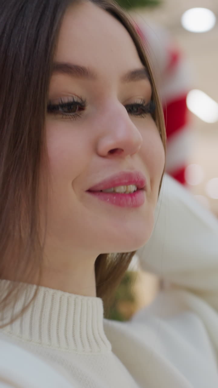 Elegant lady in white sweater posing for a photo in front of Christmas tree in mall, with blurred background showing shoppers passing by