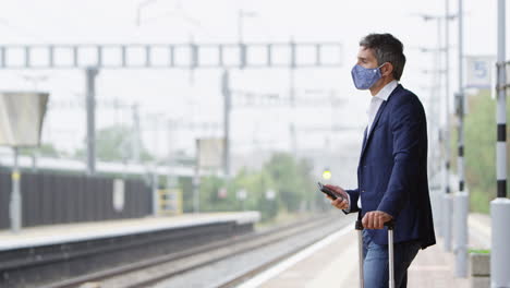 Hombre-De-Negocios-En-Plataforma-Ferroviaria-Con-Teléfono-Móvil-Usando-Máscara-Facial-Ppe-Durante-Una-Pandemia-De-Salud