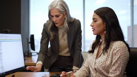 two professionals engage in a collaborative discussion while analyzing data at a workstation. the setting features large windows and urban views, emphasizing a dynamic work environment.