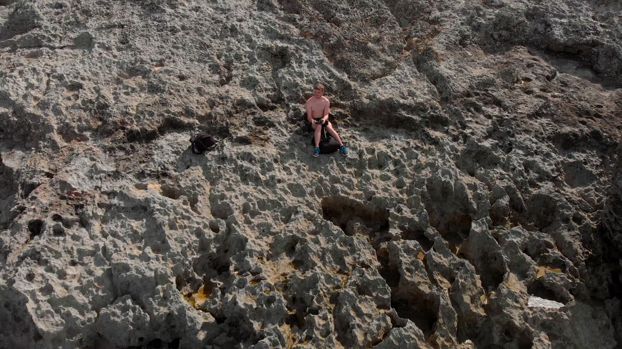 파도가 해안선에 충돌하는 것을 보여주는 석회암 절벽에 앉아 있는 남자의 반전 드론 샷