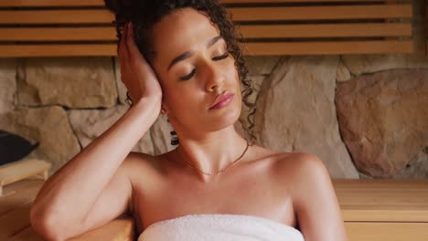 Video-of-biracial-woman-wearing-towel-relaxing-with-eyes-closed-sitting-in-sauna-room-at-health-spa