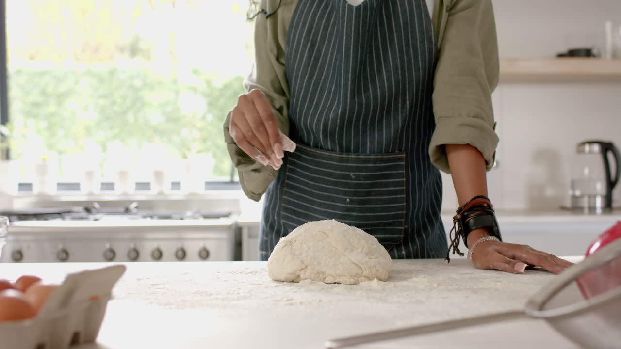 African American young woman wearing apron, making dough in kitchen