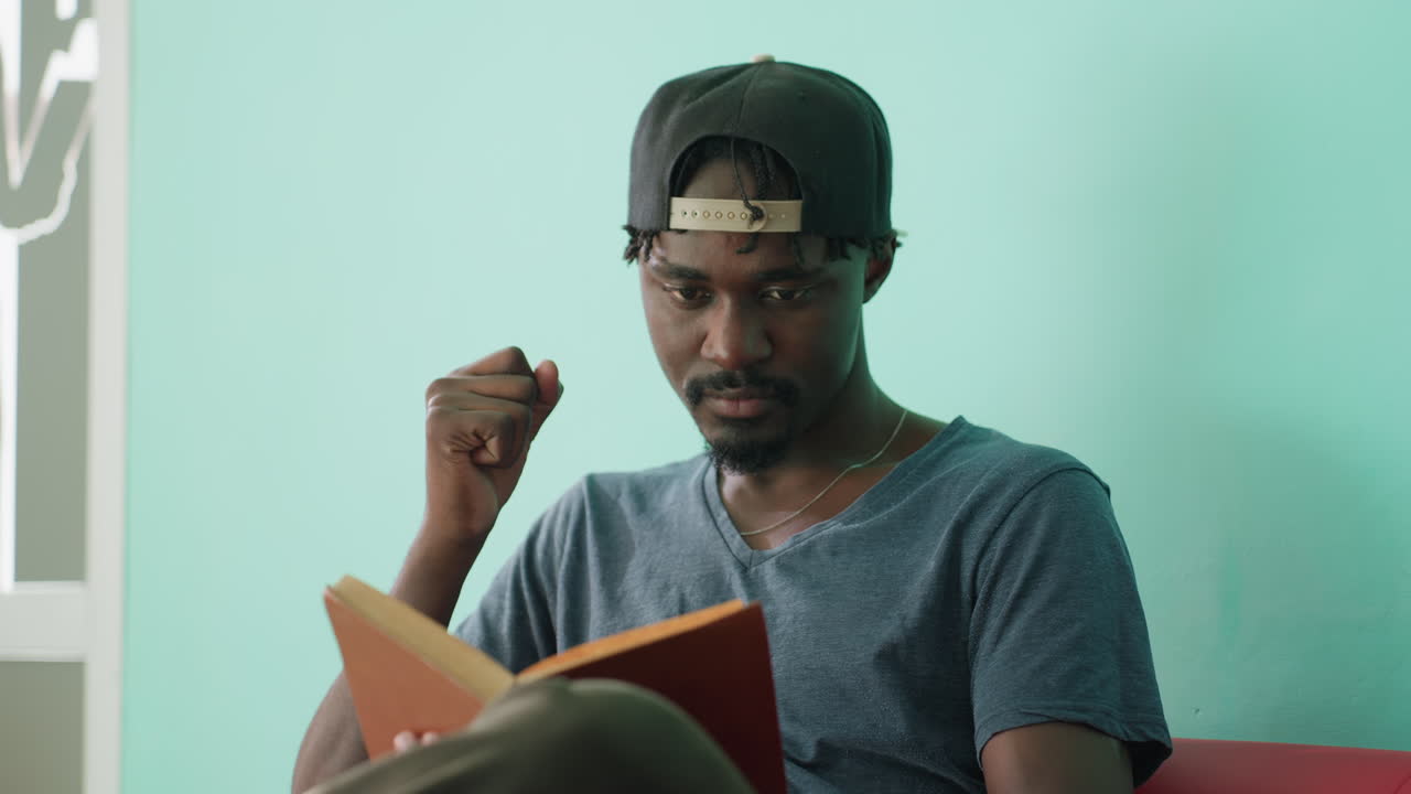 Thoughtful boy in casual shirt and cap reading brown cover book indoors with focused expression, hand near face, seated against turquoise wall with partial view of silhouette artwork in background
