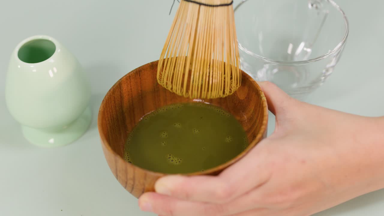 Hand vigorously whisks matcha powder in wooden bowl, bright lighting, overhead view, minimal background