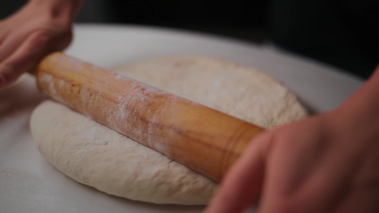 Le mani stendono l'impasto con un mattarello di legno su una superficie bianca. L'impasto è morbido e infarinato. L'azione mostra uno sforzo attento e paziente per modellare la base del pane. Adatto per cucinare, cuocere al forno. Ideale per video di ricette.