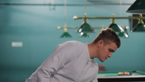 gentleman in white shirt walking in billiard room as he picks up white billiard ball and tosses it in air. focused expression, modern sports club atmosphere, green pool tables