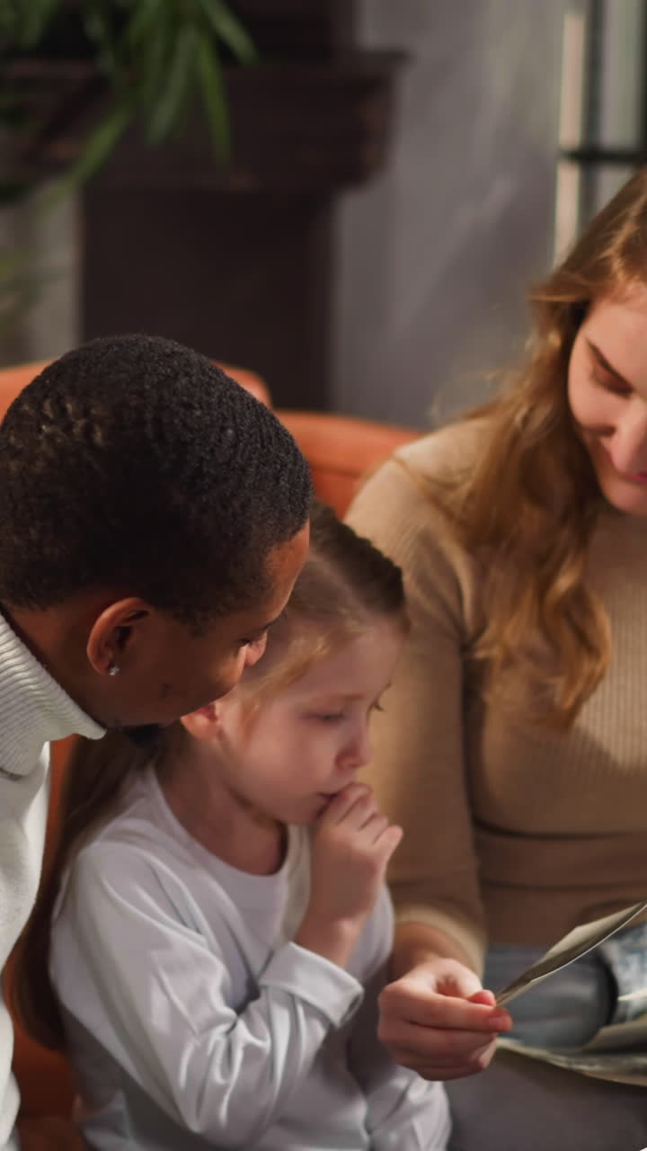 Thoughtful little girl looks at old family photos sitting with African-American father and white mother with loose hair in living room slow motion
