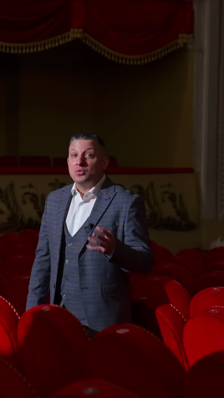 Monologue of an actor in empty theater. Professional performer in suit standing among red chairs in theatrical hall and talking on camera. Pandemic. Vertical video