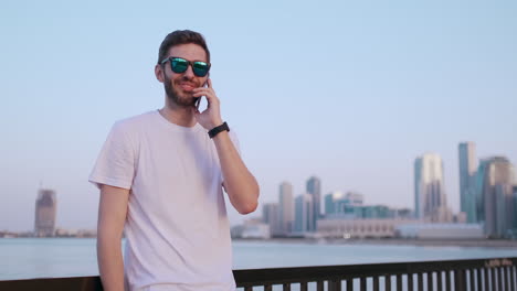 Happy-handsome-man-in-sunglasses-and-white-t-shirt-with-bristles-talking-on-the-phone-standing-on-the-waterfront-in-the-summer-against-the-city-and-buildings