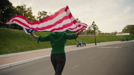 Una-Niña-De-Pelo-Corto-Verde-Con-Una-Camiseta-Verde-Y-Pantalones-Negros-Corre-Sosteniendo-Detrás-De-Sus-Hombros-La-Bandera-De-Los-Estados-Unidos-De-América,-Que-Ondea-Con-El-Viento-En-El-Parque-En-Verano.