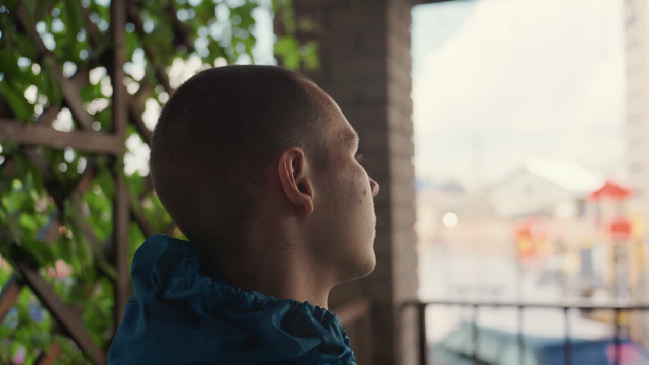 Pensive man balcony rain cinematic urban portrait, sequence of closeup head turns from profile to frontal, young male spectator soaked in drizzle wearing blue jacket, blurred city lights beyond wet