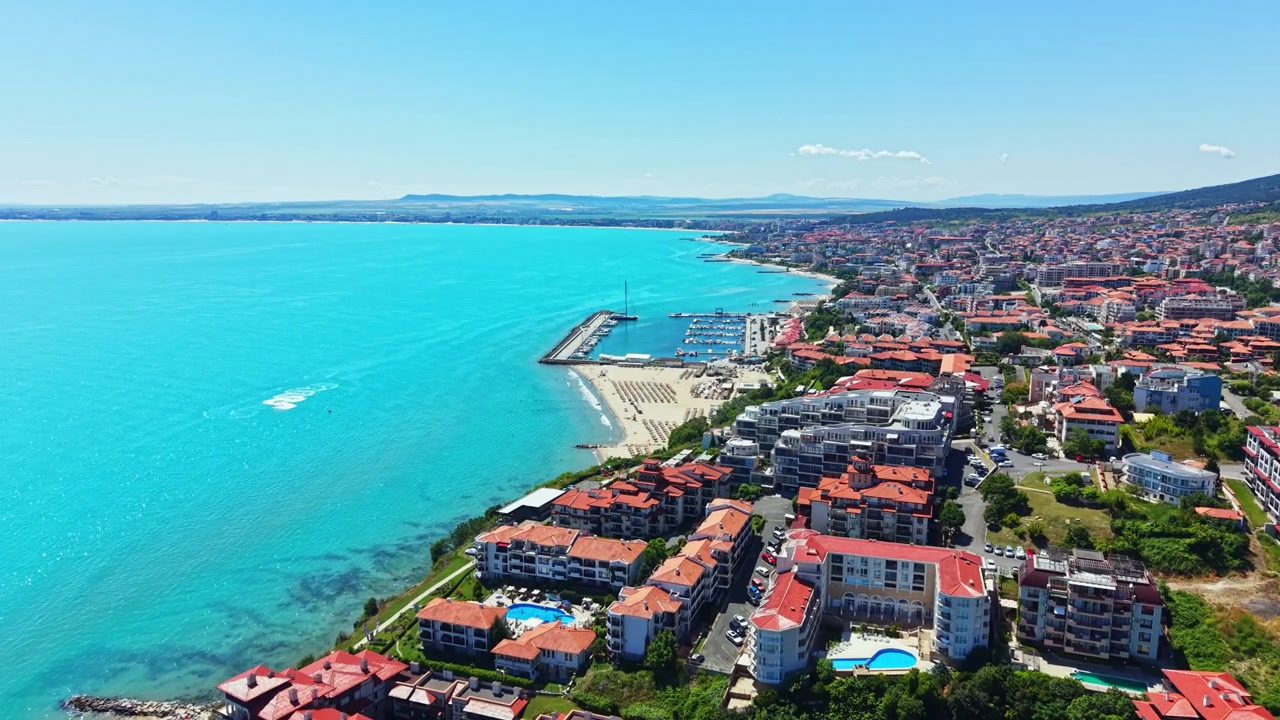 Yazın güneşli bir günde, denize kıyısı olan bir kasabanın kıyı manzarası, marinadaki tekneler ve plajdaki insanlar.