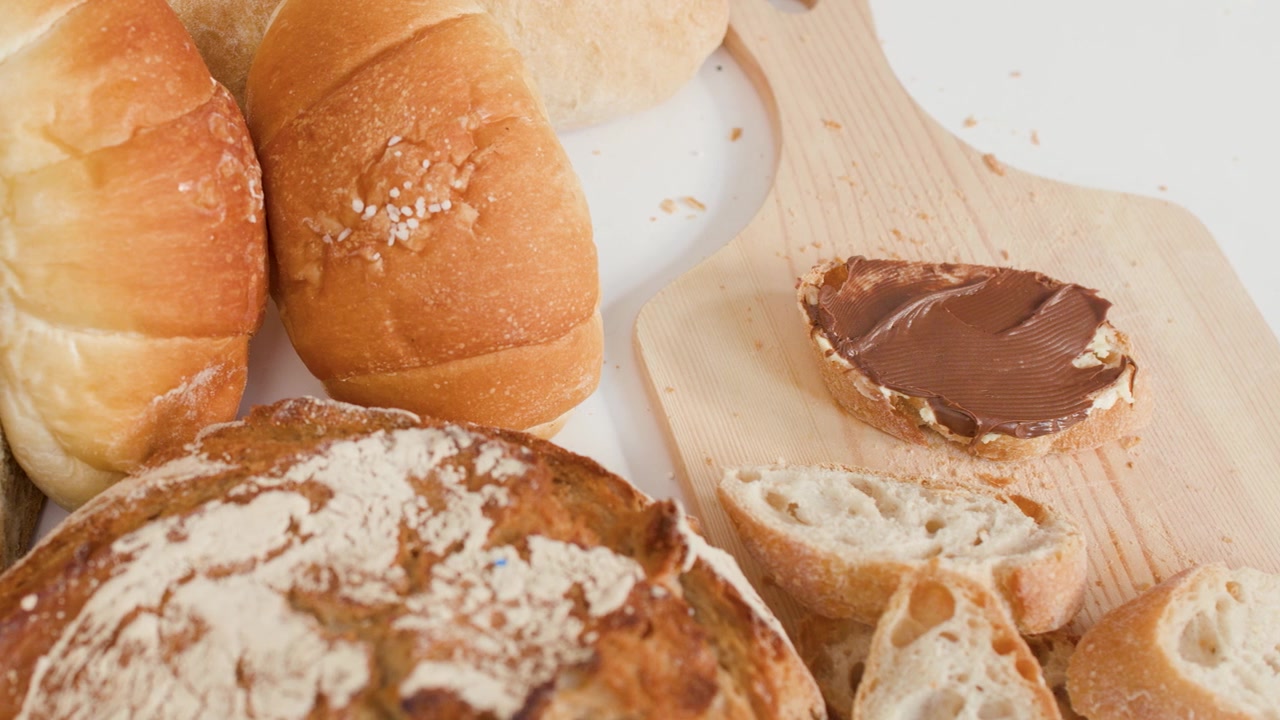 Hands use a knife to spread chocolate hazelnut cream on bread among various bakery pastries.