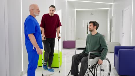 a brunette man in a green sweater sits on a wheelchair near a man who stands on crutches and talks with the doctor about their injuries and the duration of rehabilitation in a modern clinic