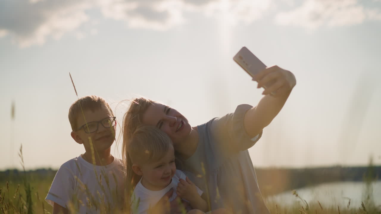 una mujer con un vestido azul se sienta en la hierba, sosteniendo un teléfono inteligente para tomar una foto de sí misma y sus dos hijos, que llevan camisas blancas, con un hermoso lago en el fondo