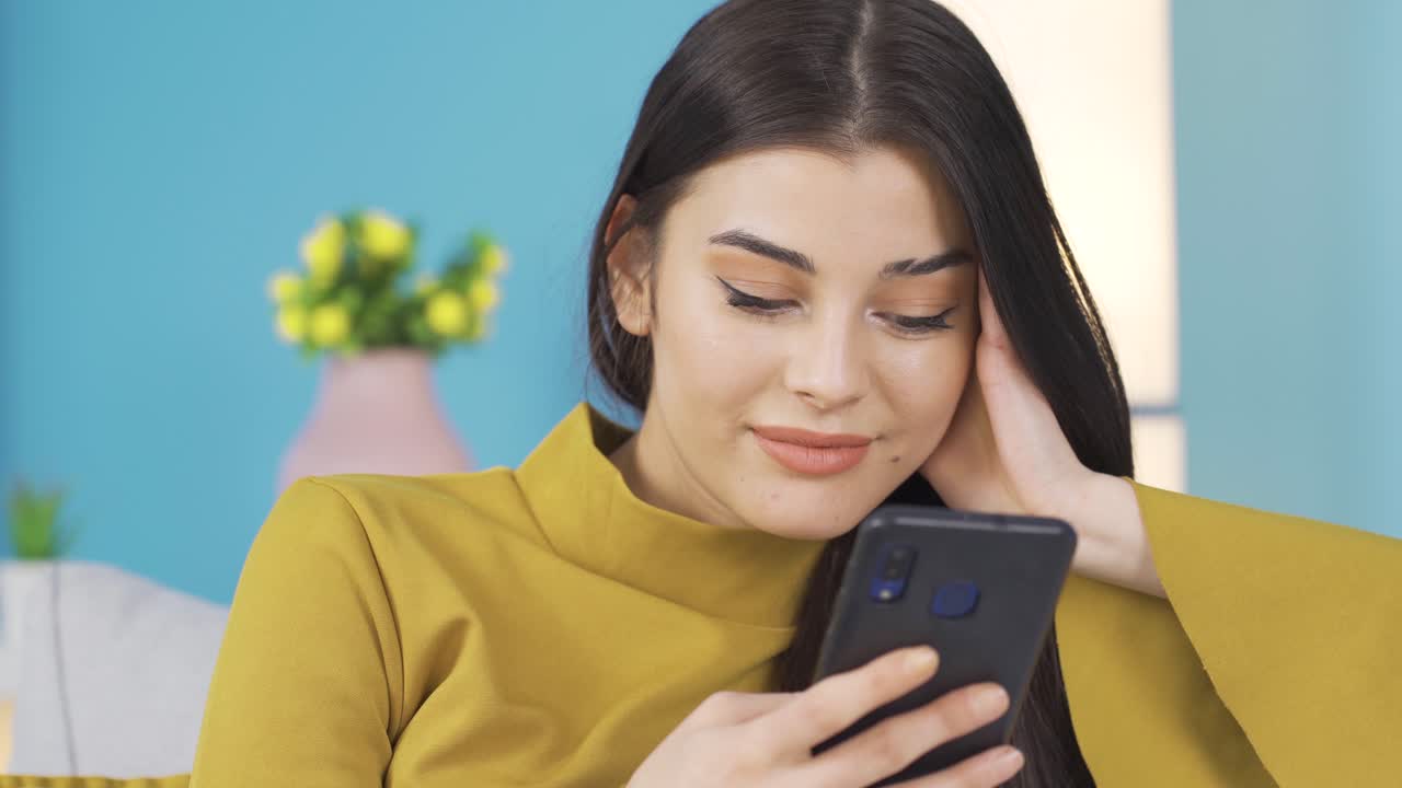 행복한 여성이 스마트폰을 사용하여 집의 소파에서 휴식을 취하고, 소셜 미디어 게시물을 보고, 온라인 쇼핑을 즐기고 있습니다.