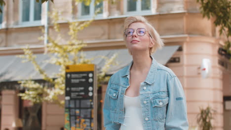 happy caucasian young woman tourist traveler in glasses walking while looking around on city street