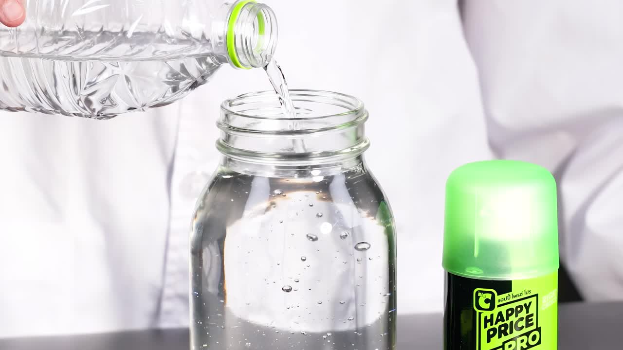 A person pours water from a bottle into a glass jar, with a green-lidded container beside it.
