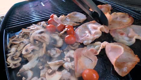 Drehen-Von-Speck,-Pilzen-Und-Tomaten-Mit-Einer-Zange-Auf-Einem-Heißen,-Rauchigen-Grill