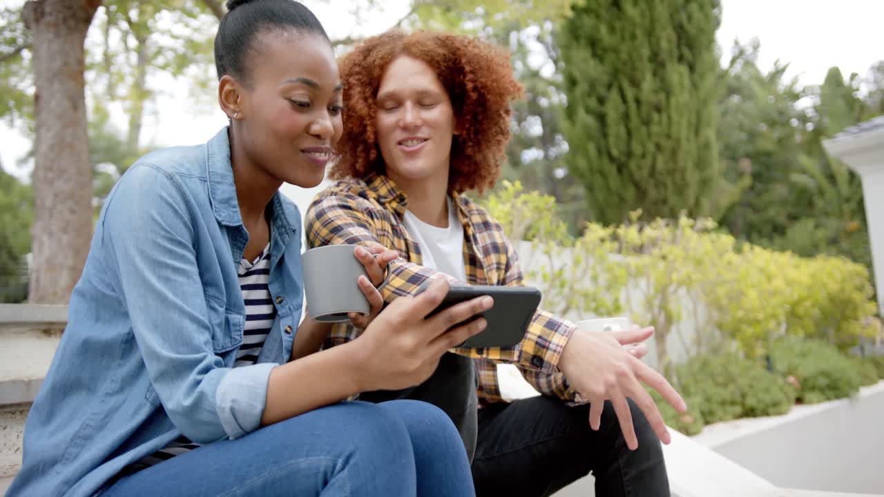 Happy diverse couple sitting on steps in garden with coffees, using smartphone, talking, slow motion