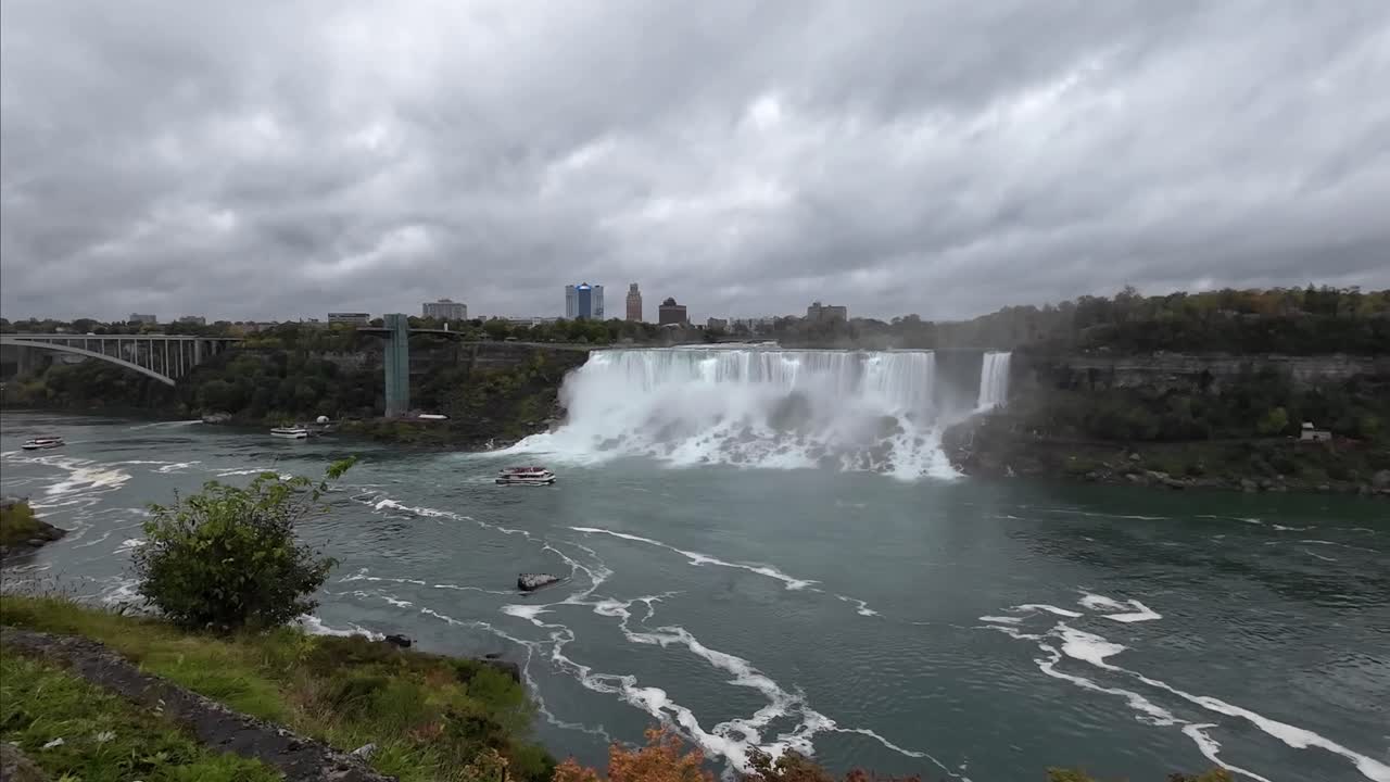 Wodospad Niagara od strony kanadyjskiej, z widokiem na wodospady po stronie amerykańskiej w pochmurny dzień, z łodziami pływającymi po wodzie przed wodospadem.
