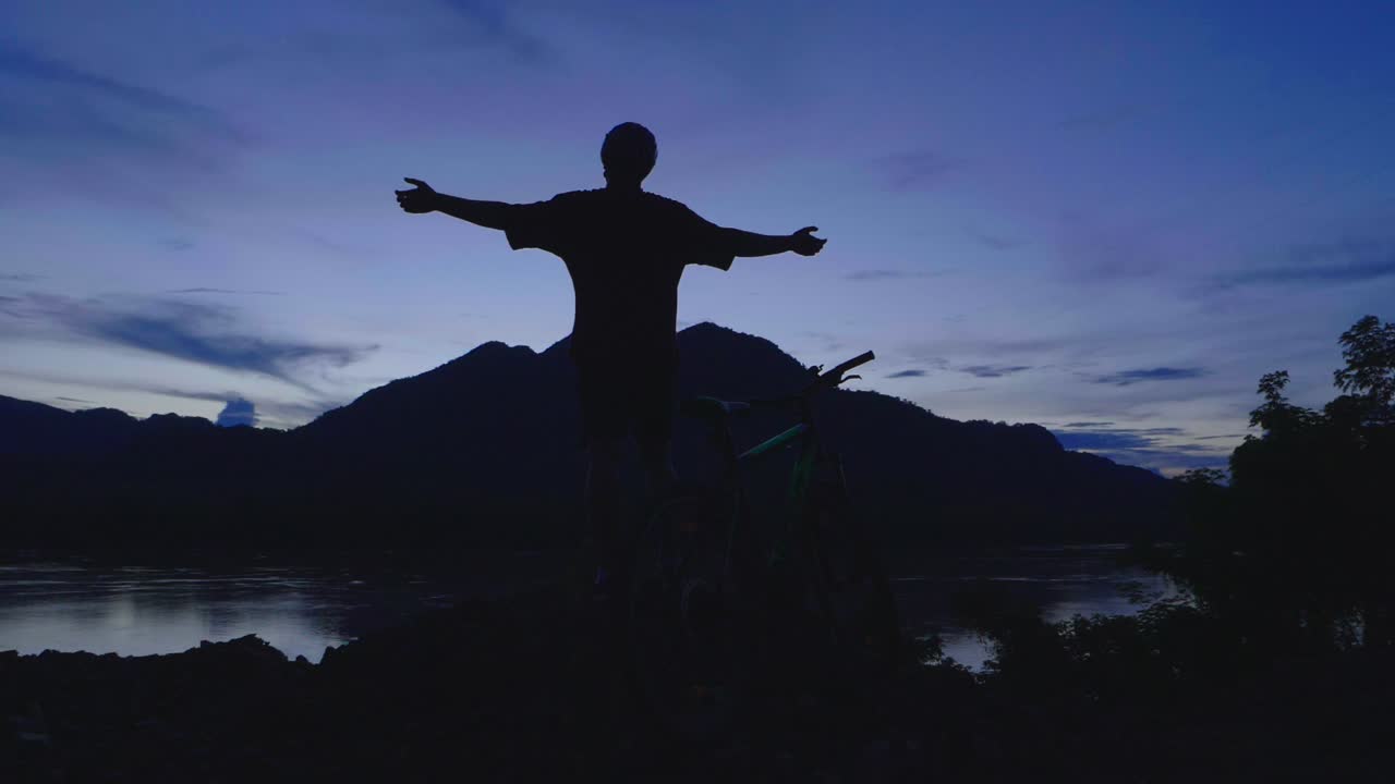 Silhouette of a Man with Arms Outstretched at Sunset over River and Mountains with Bike