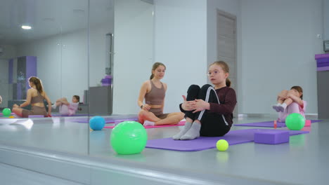 children in a bright gym perform exercises on mats, rolling back with hands clasped on knees, under the watch of an instructor visible in a reflective mirror