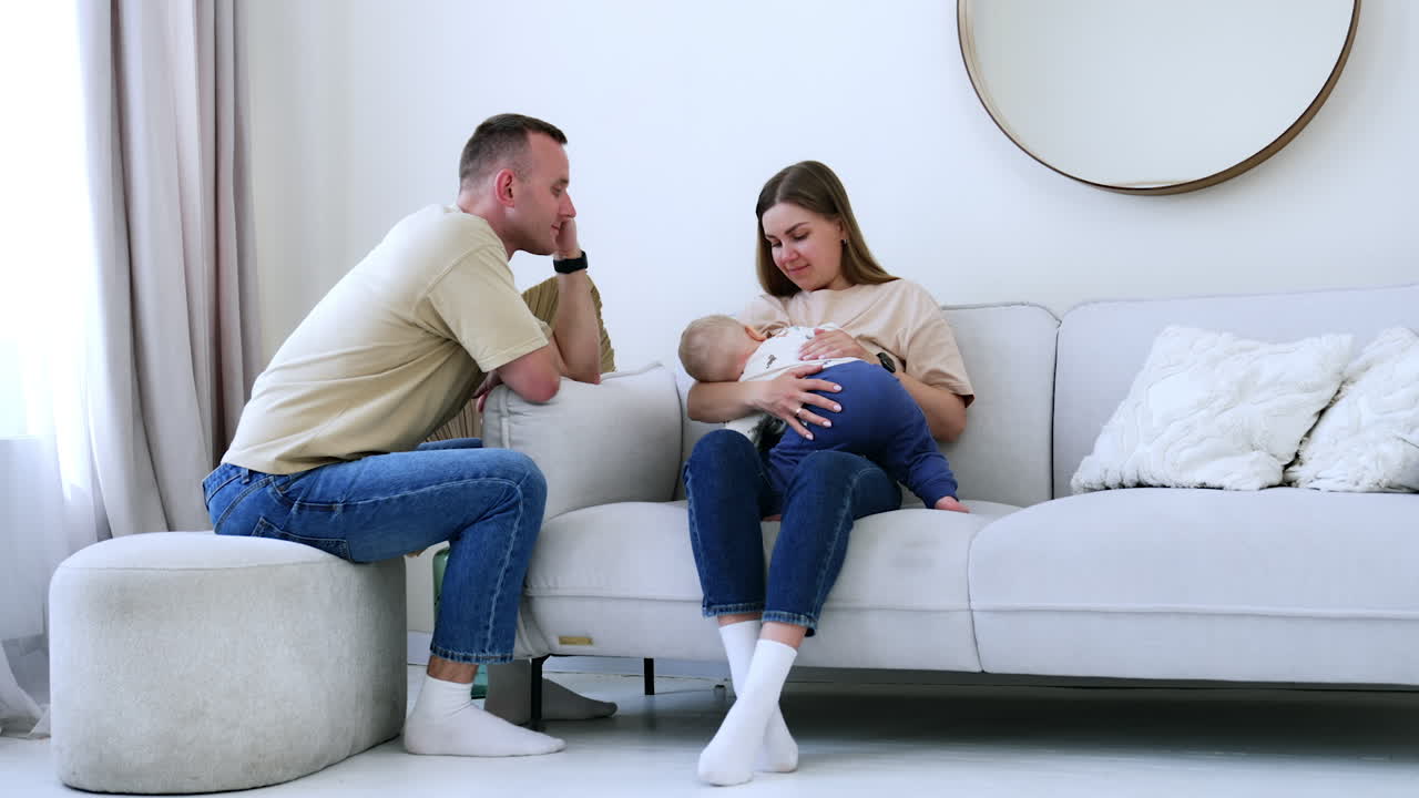 Caucasian mother sitting on the sofa breastfeeds her little baby. Man sits beside looking at his child.