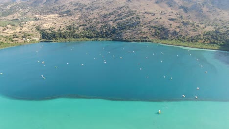 Drone-view-in-Greece-flying-over-a-light-and-dark-blue-lake-with-small-boats-and-surrounded-by-green-mountain-on-a-sunny-day-in-Crete