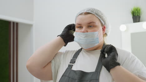beautician wearing disposable shower cap and black gloves adjusts medical mask in clean, well-lit salon with blurred background including indoor plant and mirror with lights