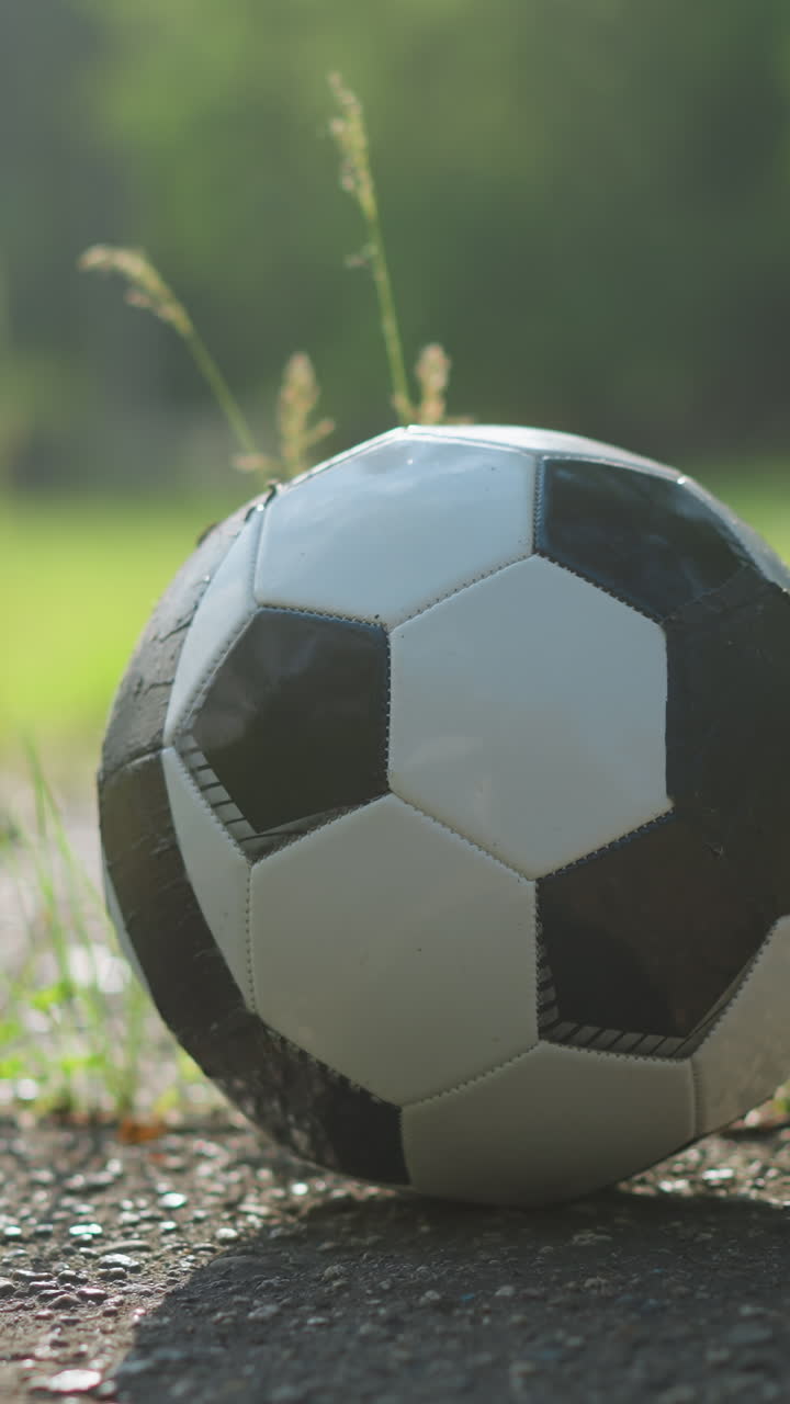 A blurred view of someone wearing brown shorts walking towards a soccer ball placed on a paved surface, with a lush grassy field in the background