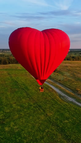 hot air balloon flying over field. romantic red aerostat in the heart shape moving slowly in the air. fantastic view on the airship in summer. vertical video