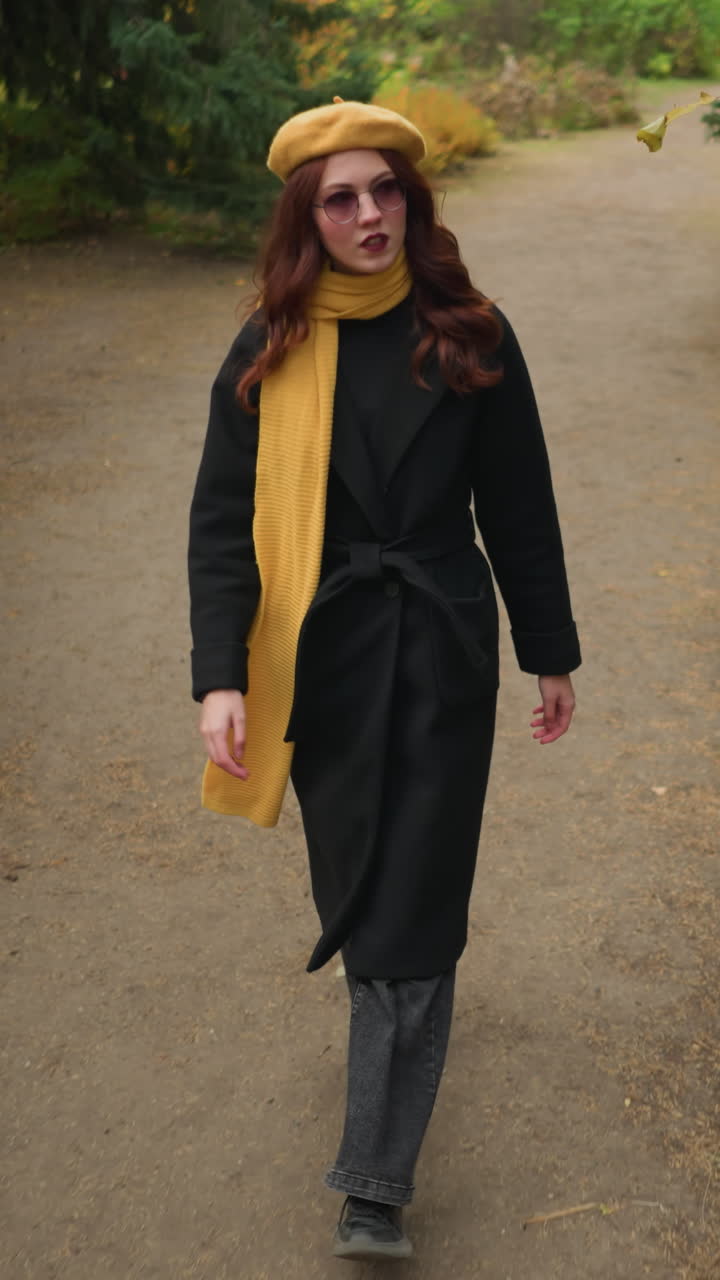 Una donna sicura di sé in cappotto nero, con un basco giallo e una sciarpa abbinata, alza lo sguardo e concentra la sua attenzione su qualcosa di intrigante sullo sfondo di un parco sereno.