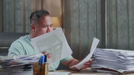 close up of fat asian man yawning and putting hand on cheek while working hard with documents at the office