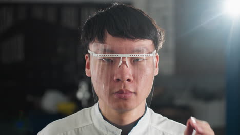 automobile engineer wearing white lab coat adjusts protective glass in automotive workshop, background features green light from machine, industrial tools, and a blurred workspace