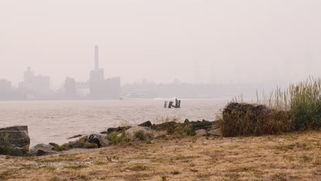 View-of-Manhattan-covered-in-smoke-from-wildfires-seen-from-park-allong-the-east-river-with-grass-and-rocks-in-the-foreground-and-ferry-crossing-river-in-the-distance