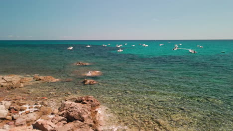 Barcos-Amarrados-En-Un-Océano-Tranquilo-Con-Costa-Rocosa-Y-Agua-Transparente-En-Un-Día-Soleado-En-Cerdeña,-Italia